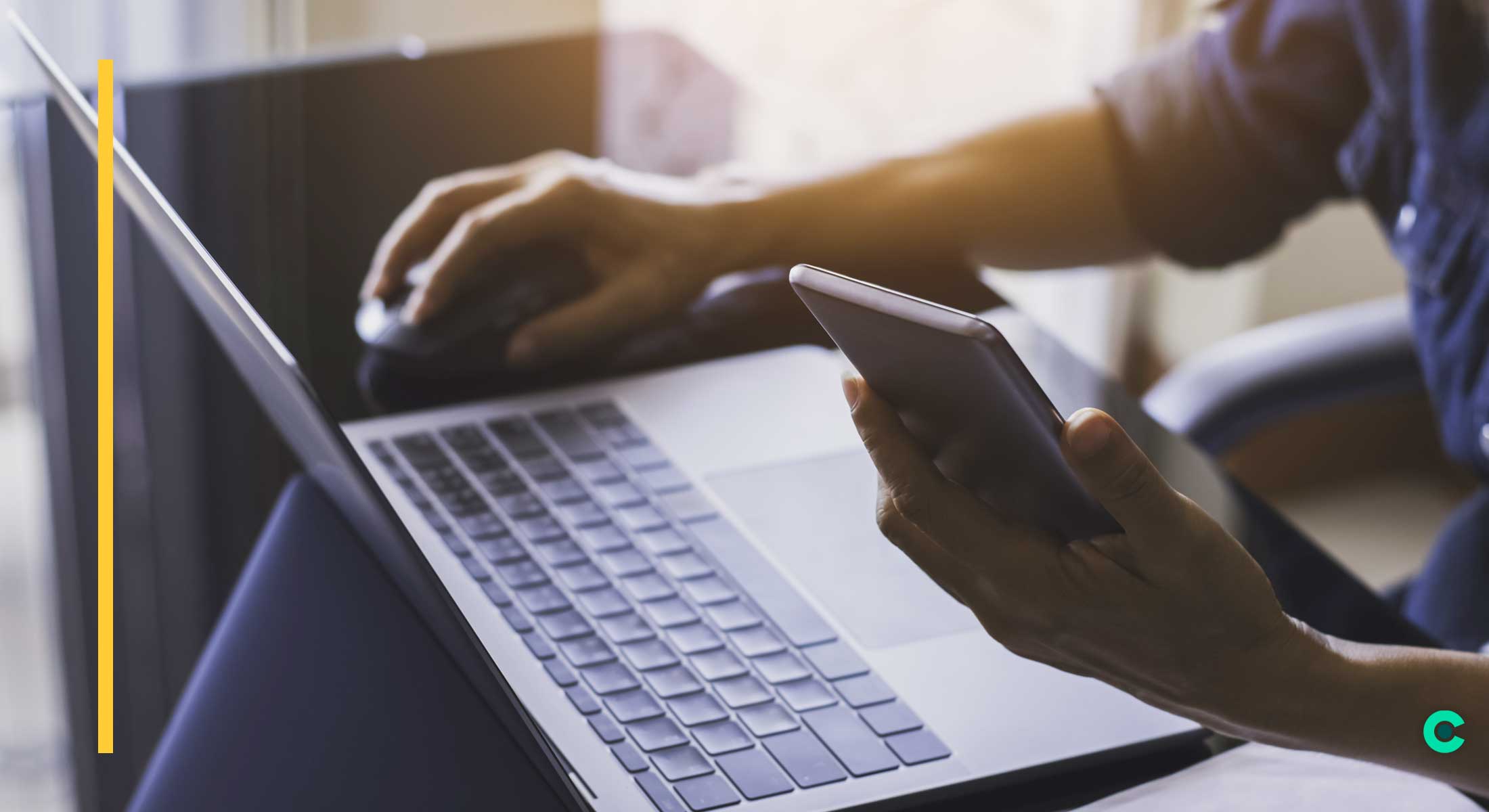 A man types on a laptop while checking his phone for emails