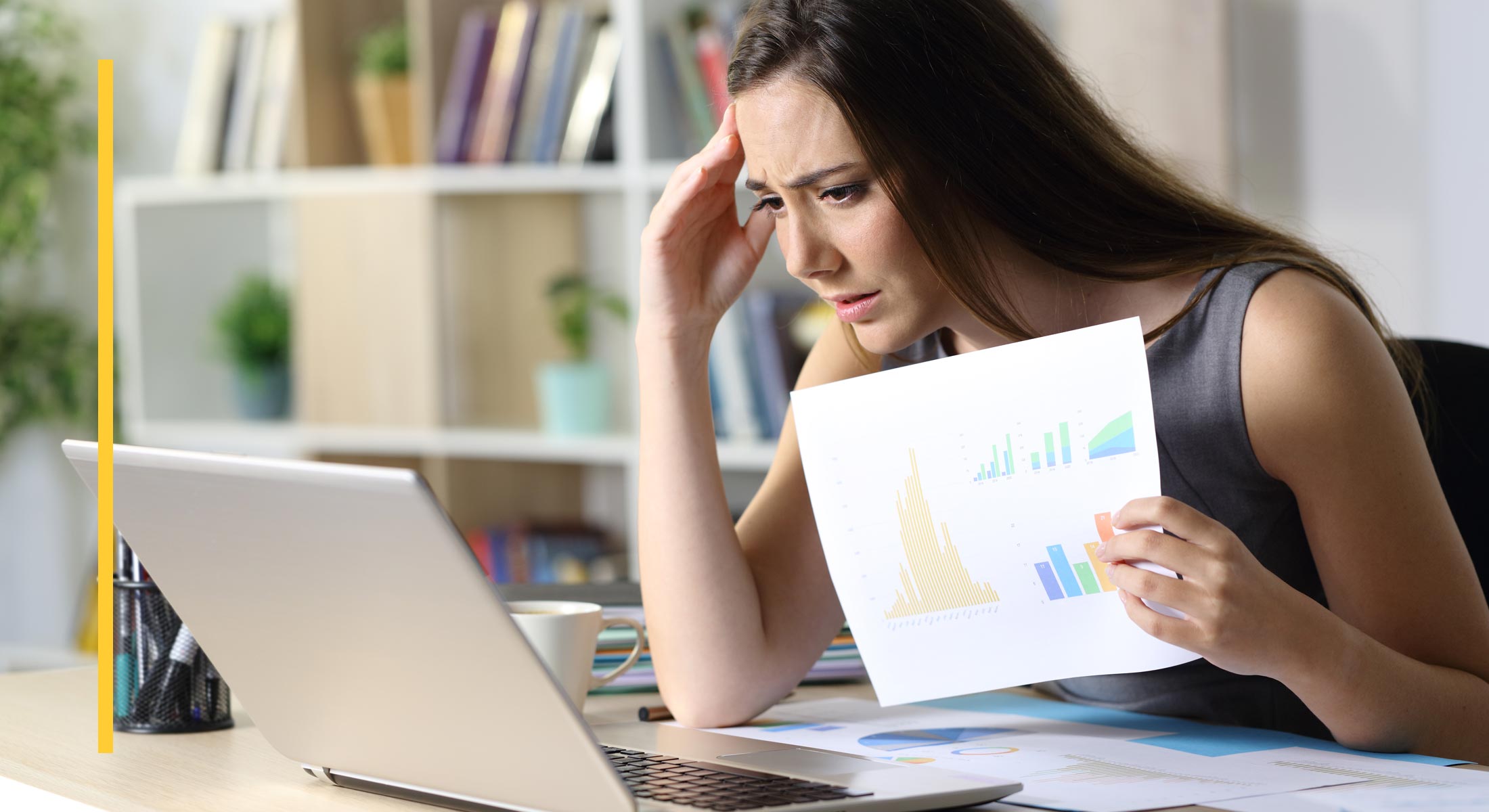 A frustrated woman holds a print-out of graphs while looking at her laptop