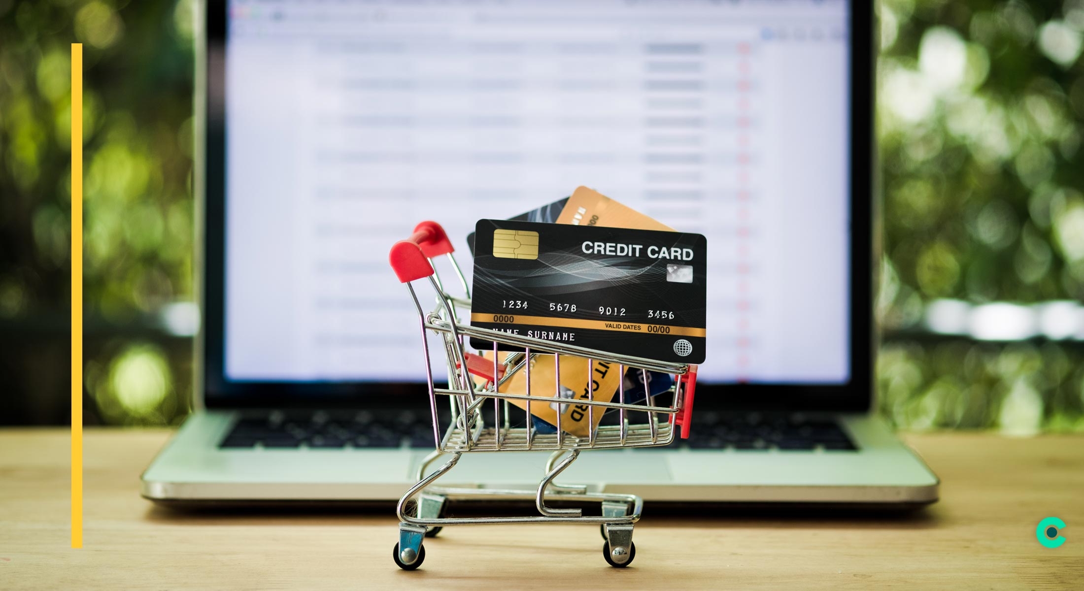 A tiny grocery cart filled with credit cards sits in front of a laptop displaying a checkout screen