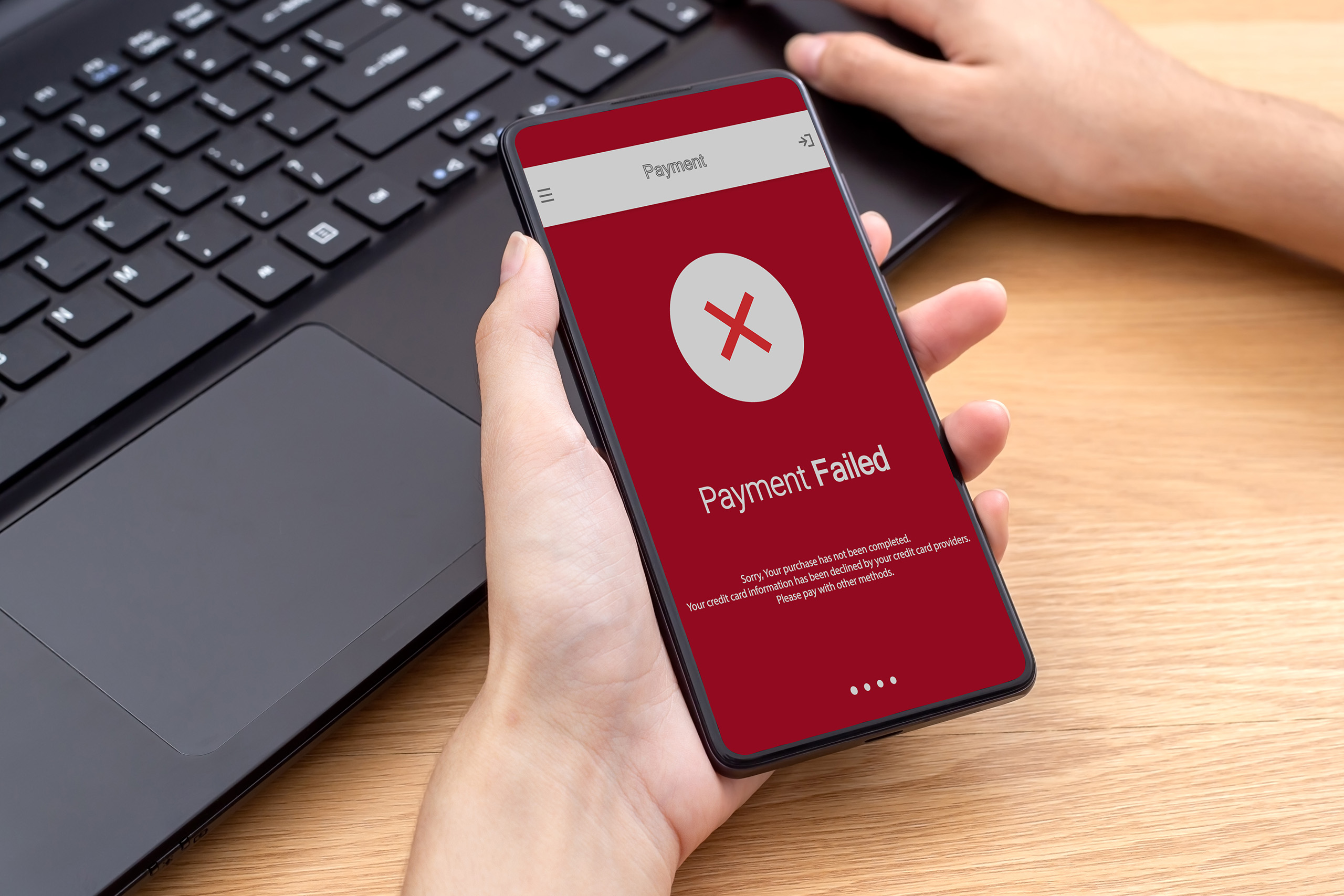 A person holding a smartphone displaying a "Payment Failed" message on a red screen, with a laptop keyboard in the background