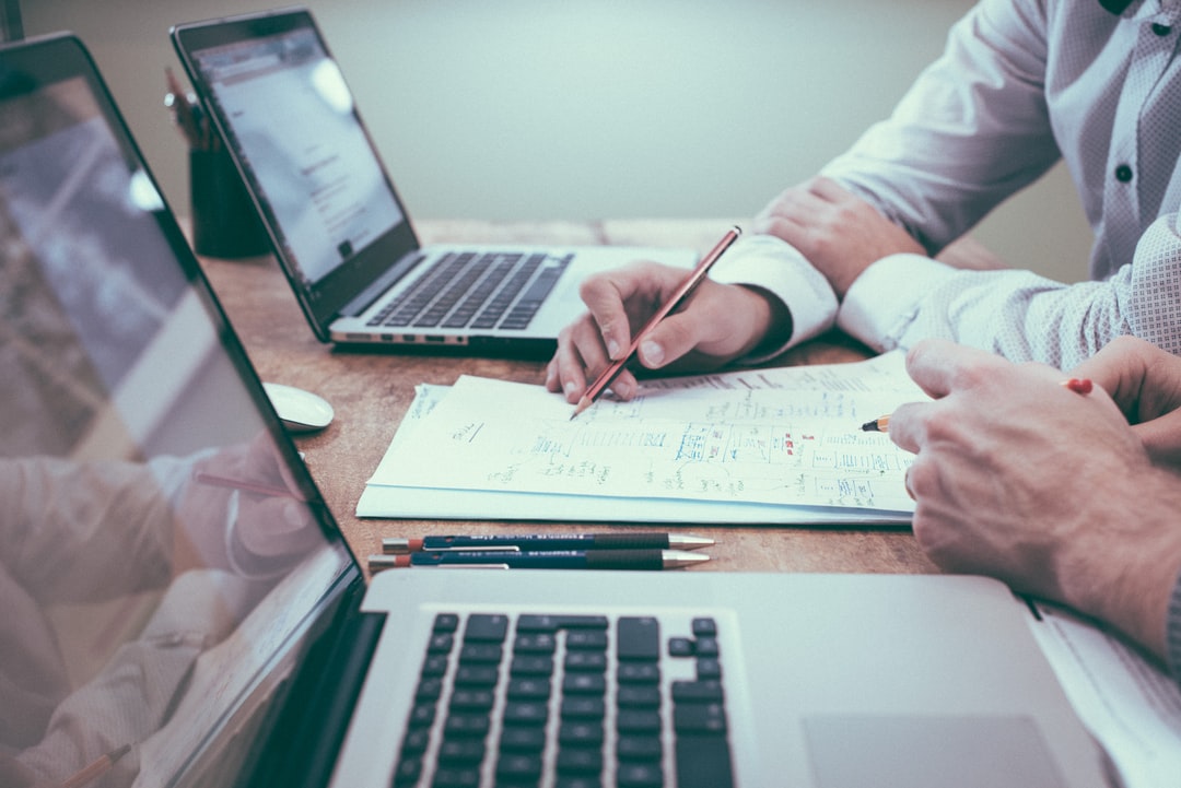 This image shows two people working together at a desk with laptops and documents.