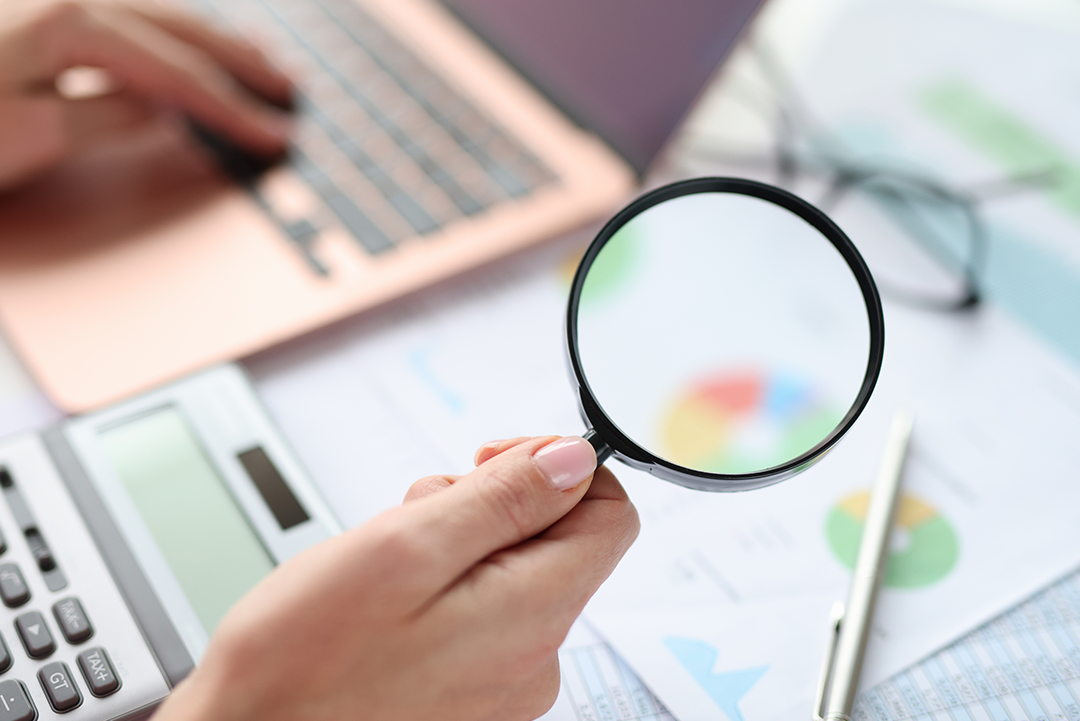 The image suggests an auditing process, with a person using a magnifying glass to examine documents, alongside a laptop, calculator, and pen.